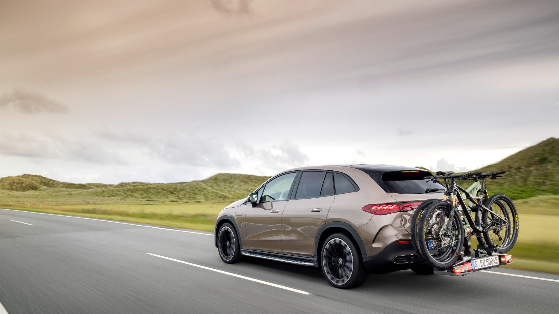 New Mercedes-Benz EQE SUV in a driving shot, from rear passenger side with bikes attached and countryside backdrop