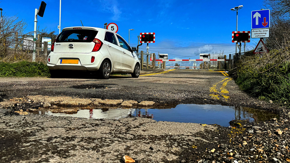 Pothole filled with water in foreground with Kia Picanto at a railway crossing in the background, bright blue skies
