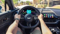 Jack Rix's arms holding the steering wheel of an Aston Martin DB7 that features Apple Carplay Ultra integration on the driver display
