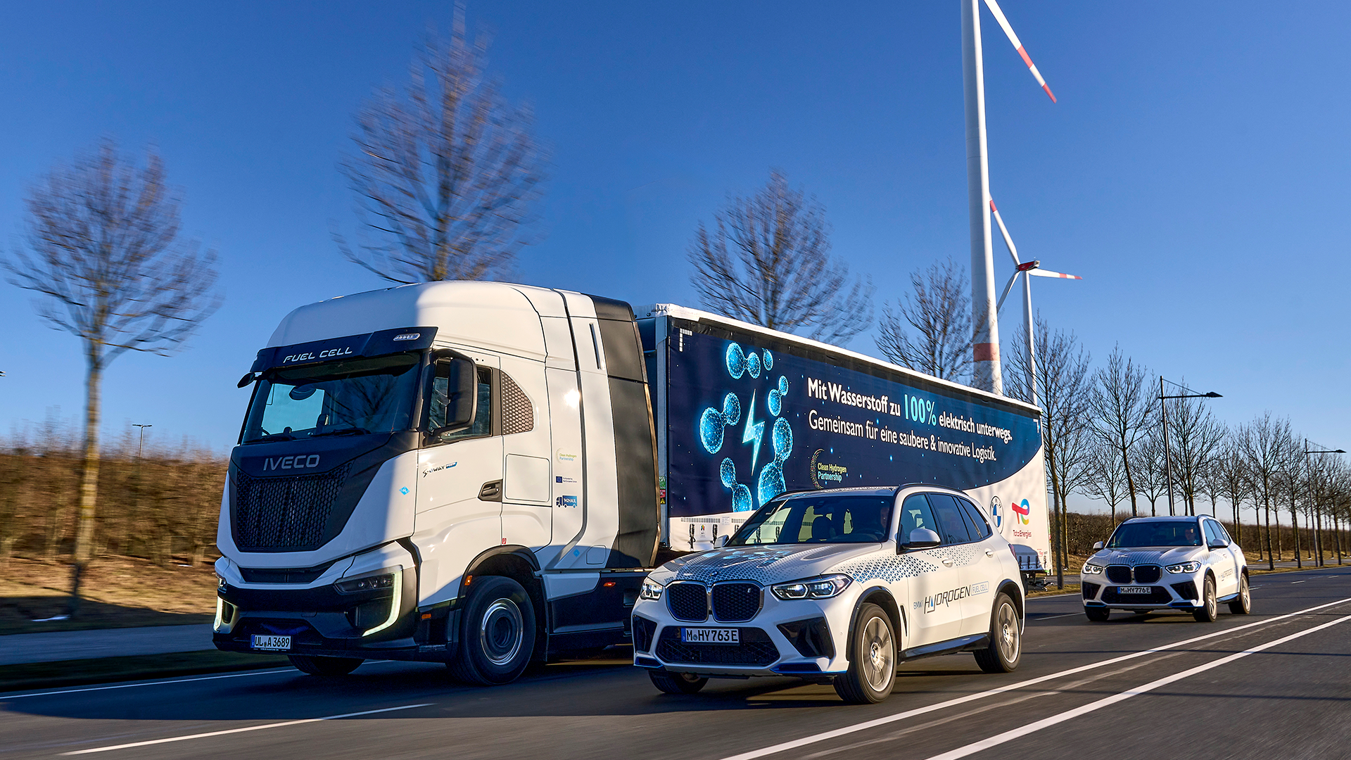 BMW iX5 hydrogen-fuelled car, alongside a truck with a fuel cell, passing wind turbines
