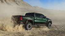 A green Ram TRX driving on a sandy desert terrain, kicking up dust under a blue sky.