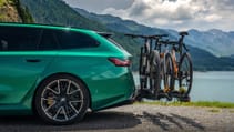 A green car with a bike rack attached to the back, parked near a lake and mountains.