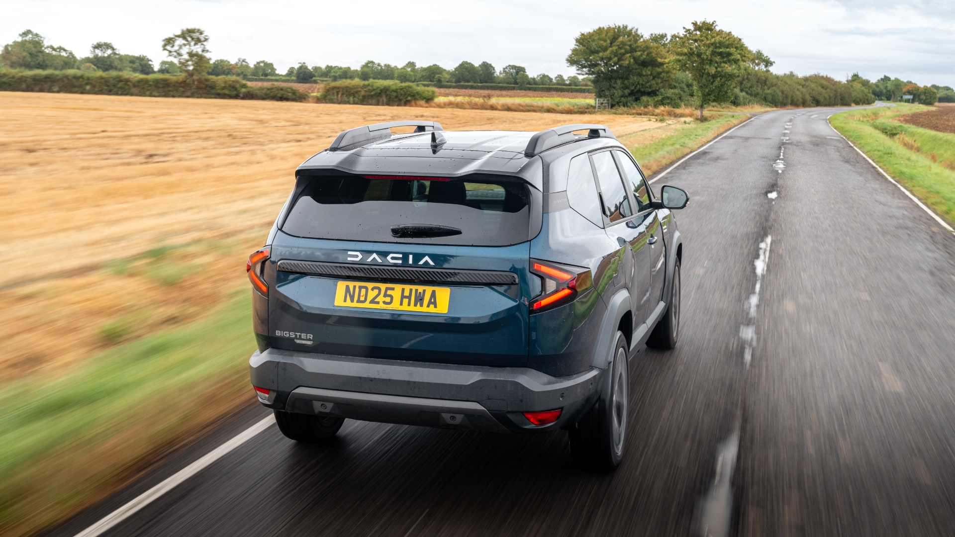 Dacia Bigster driving on a road next to a field, pictured from behind.