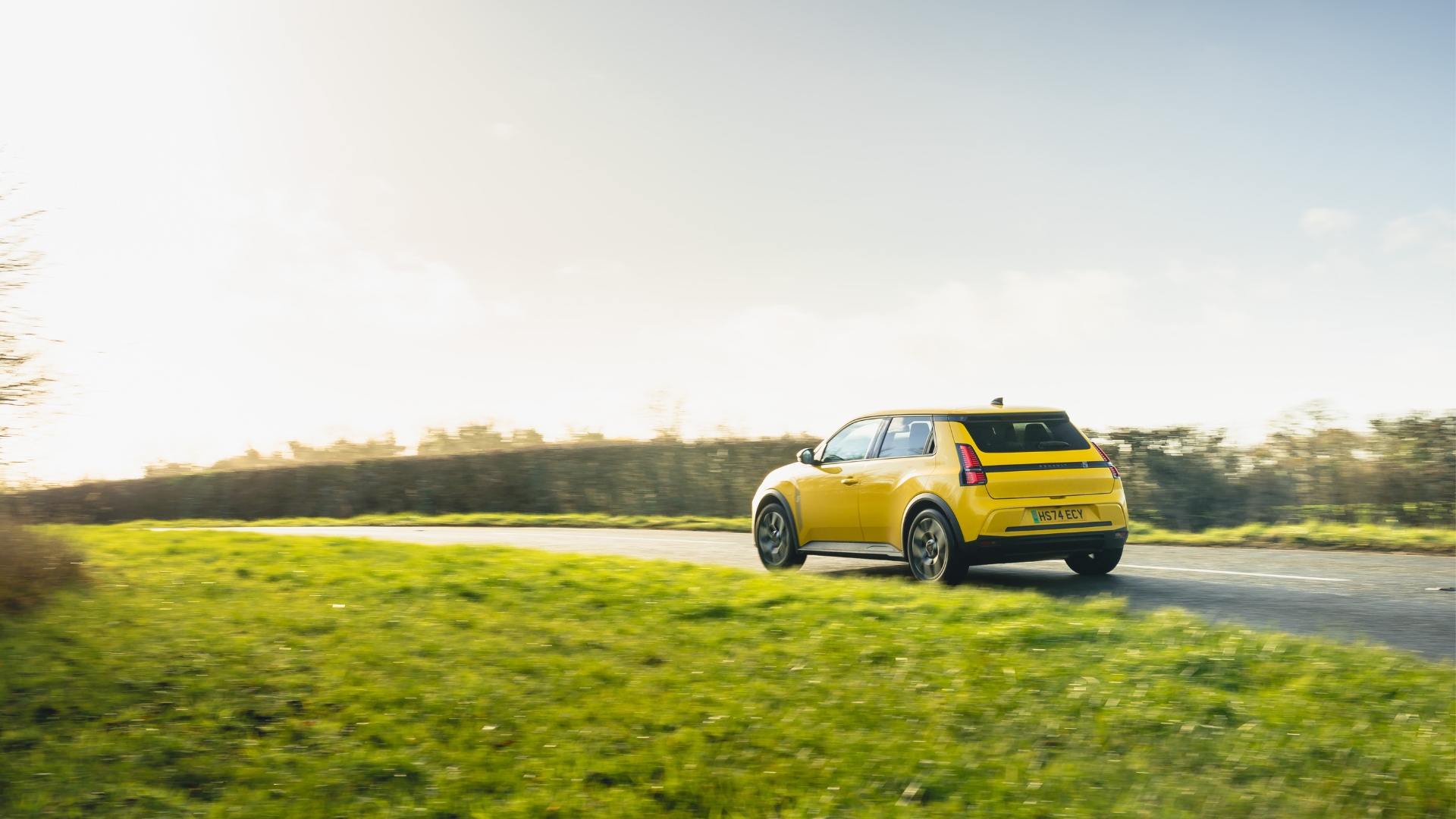 A yellow Renault 5 drives along a winding road through a grassy, green landscape.