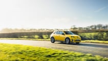 A yellow Renault 5 drives along a winding road through a grassy, green landscape.