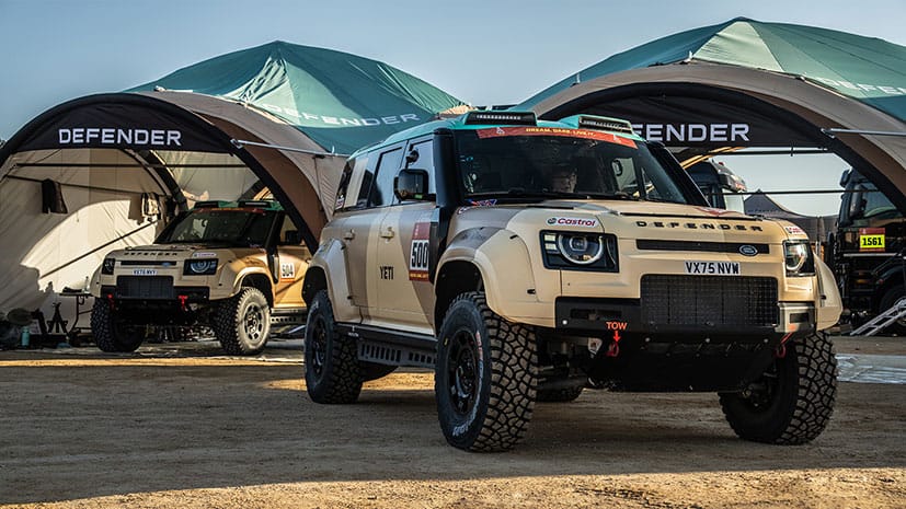 Two tan off-road Defender Dakar D7XR vehicles parked on sandy terrain under green and white tents labeled 'Defender'