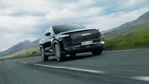A sleek, black, hybrid JAECOO 7 SUV with a prominent grille cruises down an open road, surrounded by grassy hills and a cloudy sky