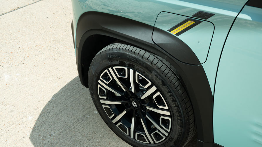 Close-up of a pastel blue Renault 4 E-Tech electric car's front left wheel and wing on a concrete surface. The black wheel has a sleek design, with white accents