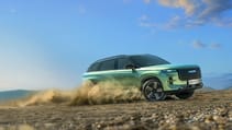 A sleek, green, hybrid JAECOO 7 SUV speeds down a dusty country road lined with trees. The sky is partly cloudy