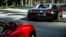 Two Bugatti Veyrons driving on a quiet country road lined with tall trees, pictured from the behind