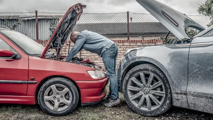 Man stands over engine bay, with two cars parked nose-to-nose. Bonnets up, he is looking to jump the battery
