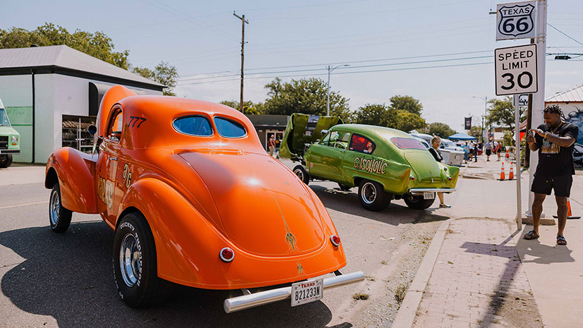 A man takes a picture of a classic car in orange on a city street, with another classic car in green in the background