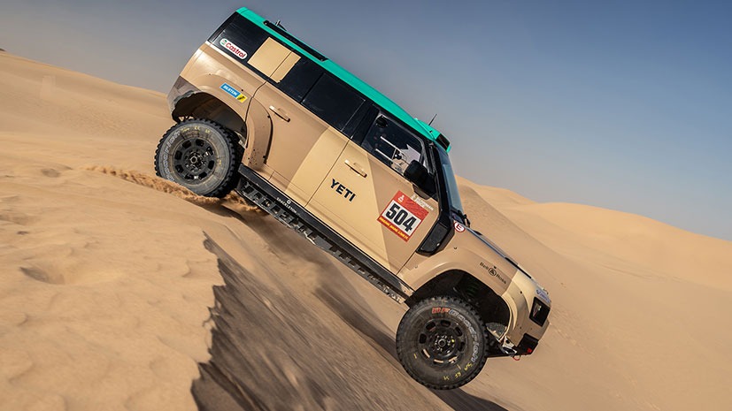 An off-road Land Rover Defender Dakar D7XR vehicle with desert rally markings ascends a steep sand dune at the 2026 Saudi Arabia event