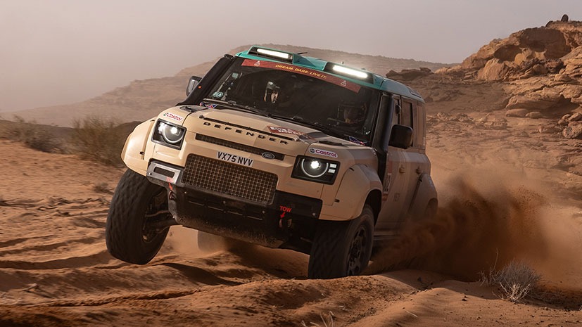 An off-road Land Rover Defender Dakar D7XR vehicle speeds through a sandy desert landscape at the 2026 Saudi Arabia event. Dust billows around it