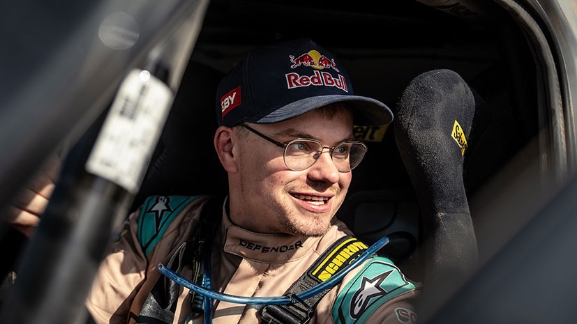 A person wearing a racing suit and cap sits in a rally car, smiling