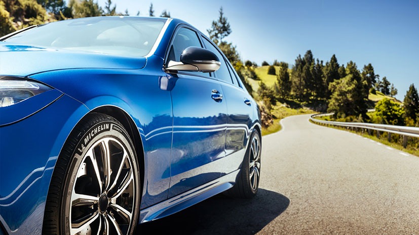 Close-up of a shiny blue car on a winding road, highlighting the Michelin tyres. The car is surrounded by trees and hills under a clear blue sky
