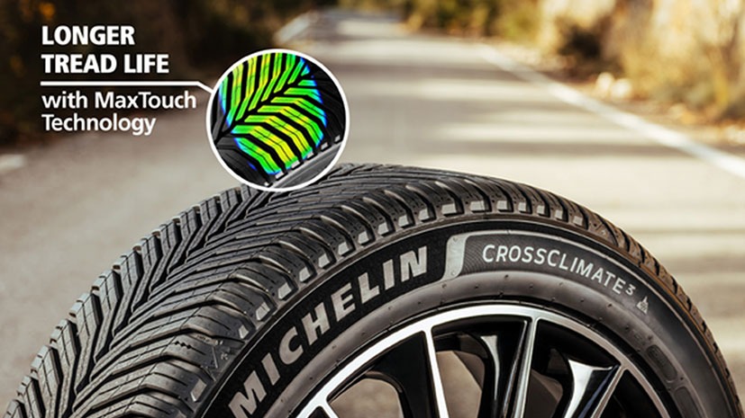 Close-up of a Michelin CrossClimate 2 tire on a road, featuring zigzag tread patterns. Overlay text highlights 'Longer tread life with MaxTouch Technology'