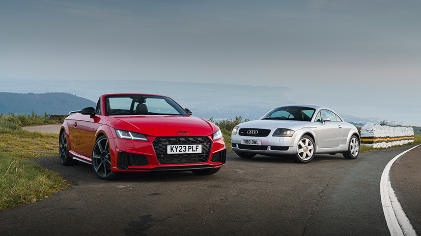 Two Audi TT RS Coupé's - one red and one silver are parked on a scenic, winding road overlooking a mountainous landscape, with the overcast sky in the background