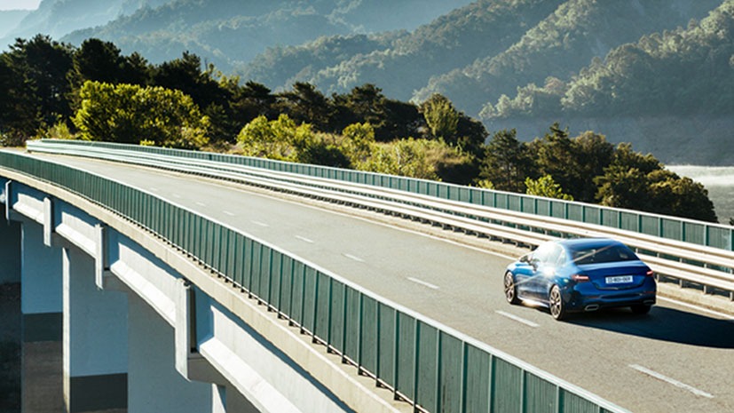 A blue car drives on a scenic highway bridge surrounded by lush greenery and distant mountains