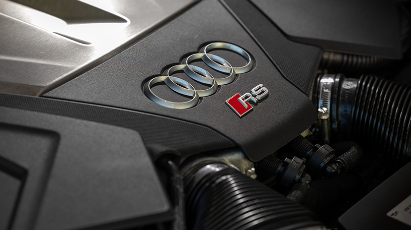 Close-up of an Audi RS car engine showcasing the Audi logo and RS badge prominently on a sleek black cover, with visible metallic components and hoses