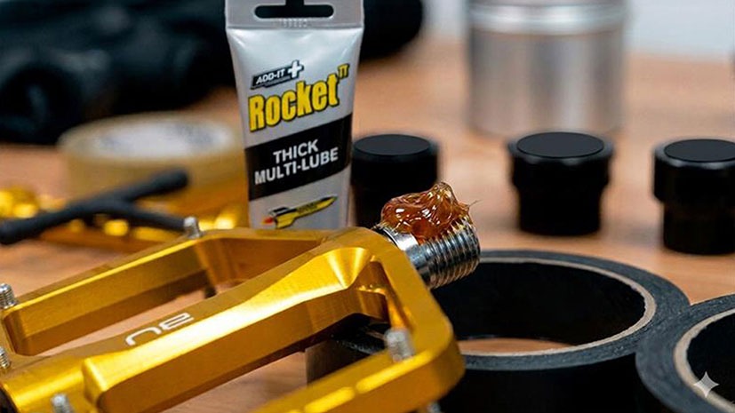Close-up of a yellow bicycle pedal with thick grease applied to the spindle. A tube of Rocket TT Thick Multi-Lube Super stands in the background, surrounded by other bike tools, on a workbench