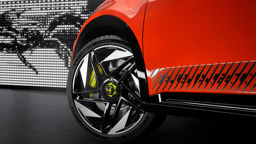 Close-up of a bright orange Abarth 600e Competizione car from a side view, featuring a sleek, modern alloy wheel with a scorpion emblem, on a glossy, black floor