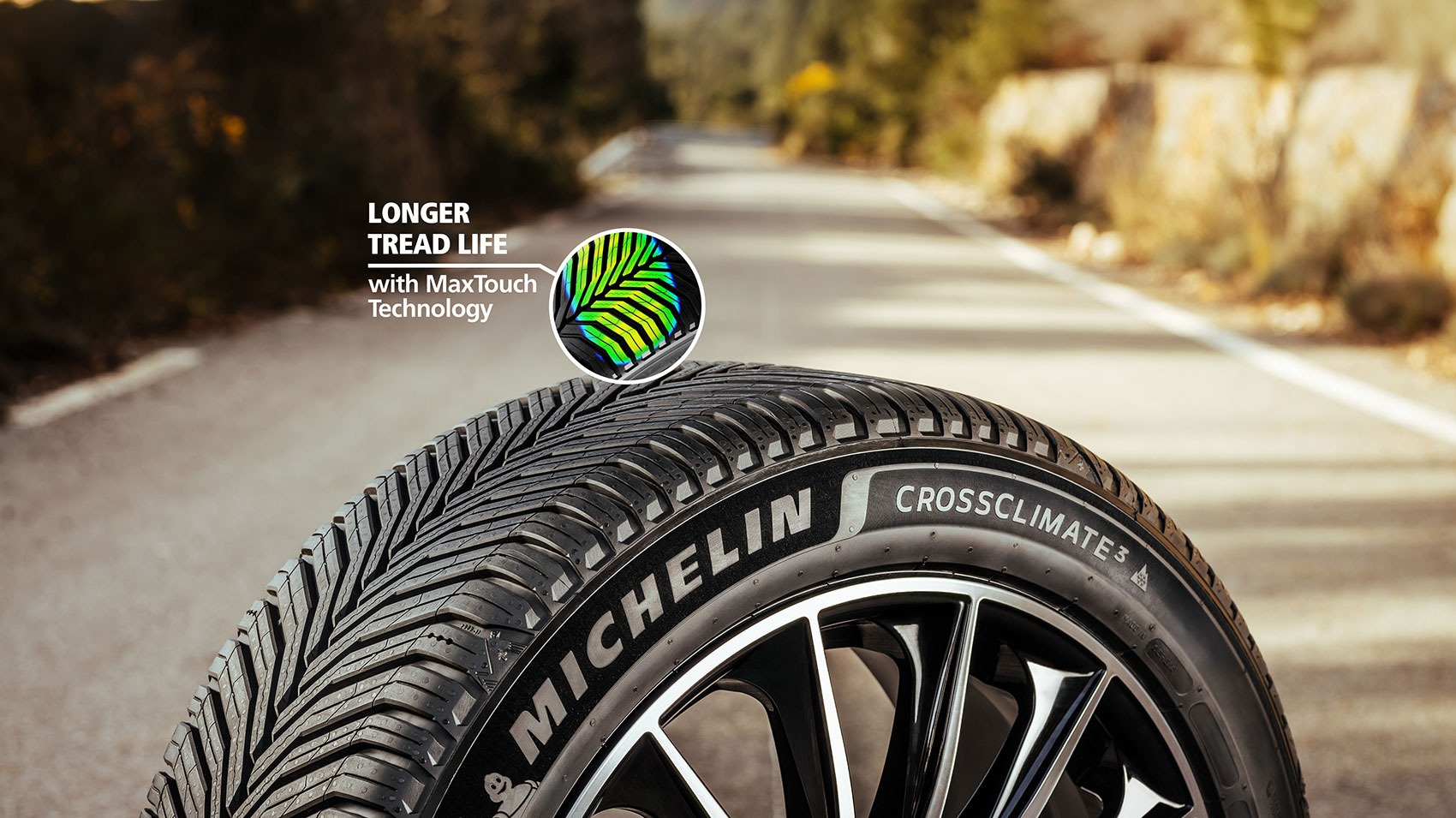 Close-up of a Michelin CrossClimate 2 tire on a road, featuring zigzag tread patterns. Overlay text highlights: Longer tread life with MaxTouch Technology