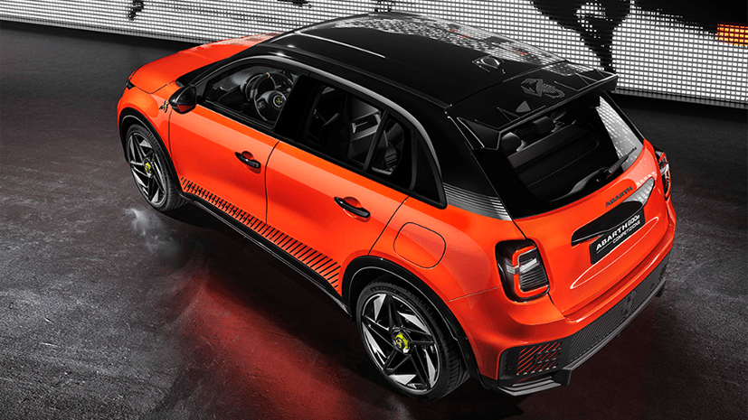 A sleek, orange, Abarth 600e Competizione car with a black roof is parked indoors. Behind it, a digital wall displays a dynamic, monochrome graphic of a scorpion