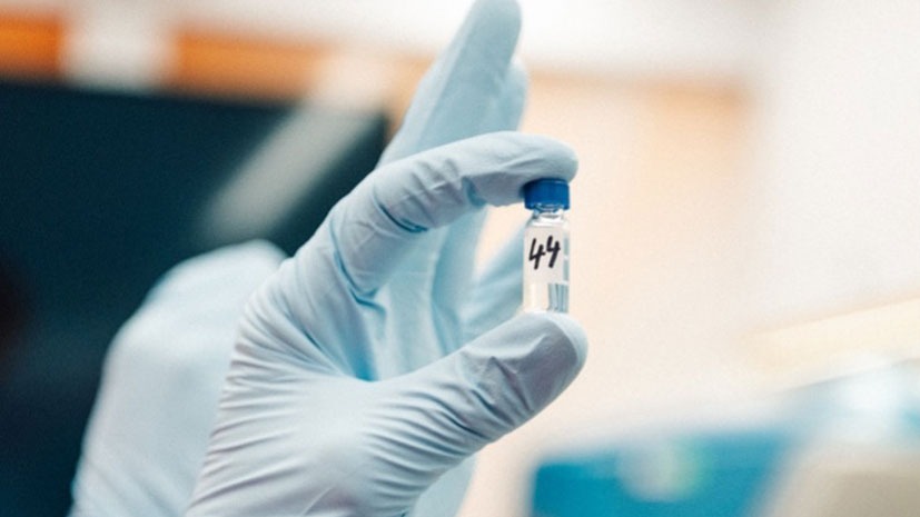 A technician's hand, wearing a powder-free blue nitrile glove, holding a small, clear glass sample vial marked with the number "44" and sealed with a blue cap