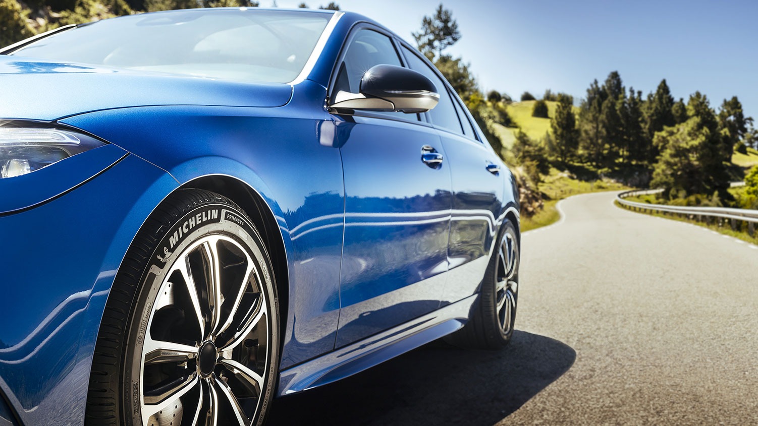 Close-up of a shiny blue car on a winding road, highlighting the Michelin tyres. The car is surrounded by trees and hills under a clear blue sky