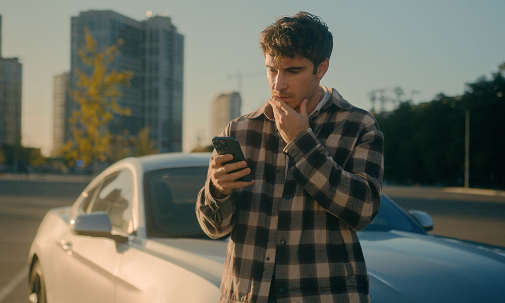 A confused driver standing next to a car looking at a mobile phone